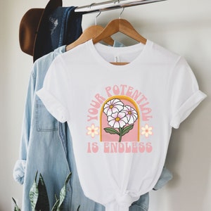 May include: White t-shirt with a pink and yellow graphic design featuring three white flowers and the text "YOUR POTENTIAL IS ENDLESS".