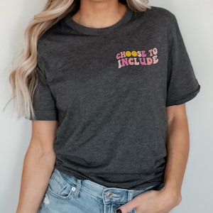 May include: A dark gray t-shirt with the text "CHOOSE TO INCLUDE" in pink and yellow retro lettering. The text is centered on the shirt and has two yellow smiley faces.