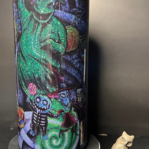 Może przedstawiać: Kubek ze stali nierdzewnej z czarno-zielonym wzorem z brokatem przedstawiającym postać Jacka Skellingtona z filmu Koszmar przed świętami. Kubek stoi na białym cokole.