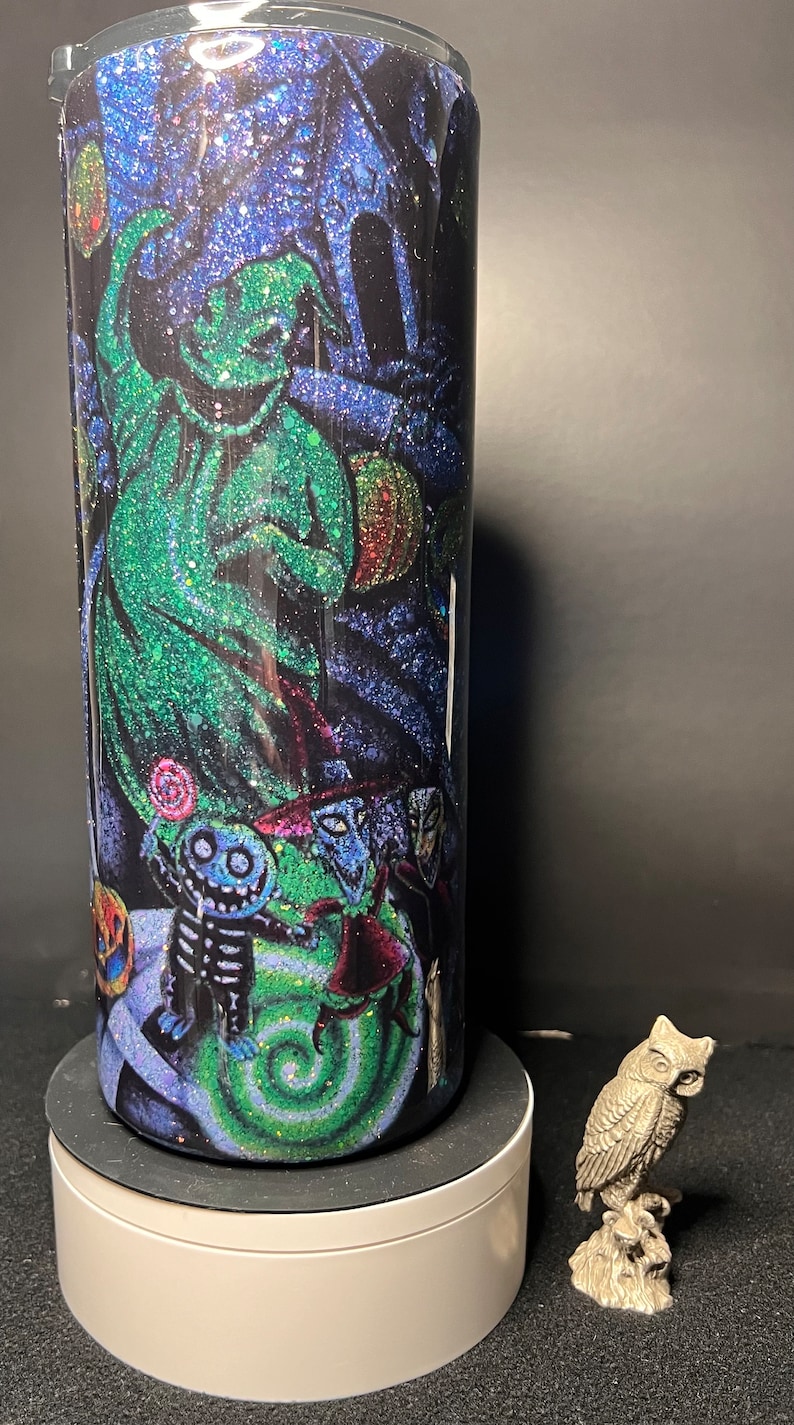 Może przedstawiać: Kubek ze stali nierdzewnej z czarno-zielonym wzorem z brokatem przedstawiającym postać Jacka Skellingtona z filmu Koszmar przed świętami. Kubek stoi na białym cokole.