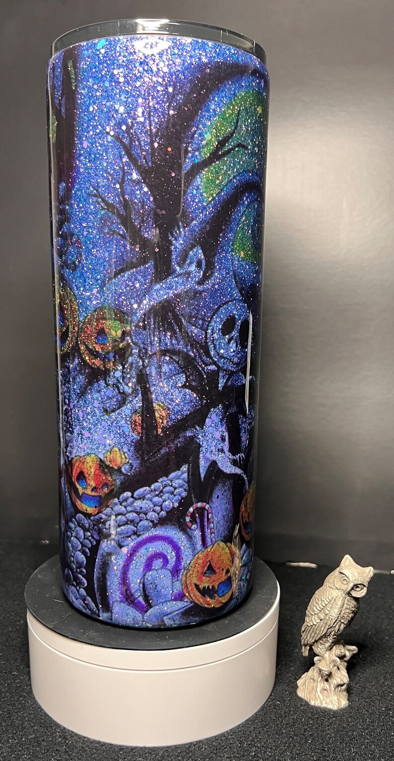 Może przedstawiać: Niebieski i zielony kubek z brokatem z motywem Halloween przedstawiającym Jacka Skellingtona z filmu Koszmar przed świętami. Wz&oacute;r zawiera dynie, laski cukrowe i straszne drzewo.