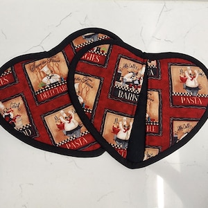 May include: A pair of heart-shaped oven mitts featuring a red background with a repeating pattern of cartoon chefs and culinary text including "DELECTABLE," "BARISTA," and "PASTA." The mitts are finished with a solid black fabric trim border.