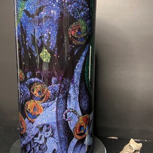 Może przedstawiać: Czarny i niebieski kubek z brokatem z motywem Halloween, przedstawiający nietoperze, dynie i laski cukrowe. Kubek stoi na białym cokole z srebrną figurką sowy na czarnym tle.