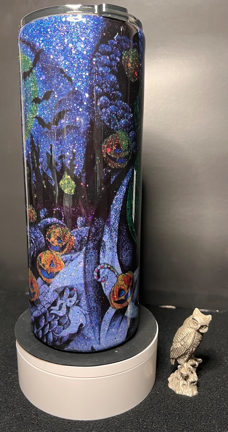 Może przedstawiać: Czarny i niebieski kubek z brokatem z motywem Halloween, przedstawiający nietoperze, dynie i laski cukrowe. Kubek stoi na białym cokole z srebrną figurką sowy na czarnym tle.