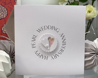 May include: A white card with the text "Happy Pearl Wedding Anniversary" in a circle around two pink hearts with a pearl in the center.