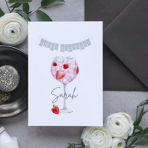 May include: A white card with a gray banner that says "Happy Birthday" with pink string. A watercolor illustration of a pink gin cocktail with strawberries and ice in a glass. The name "Sarah" is written below the glass.
