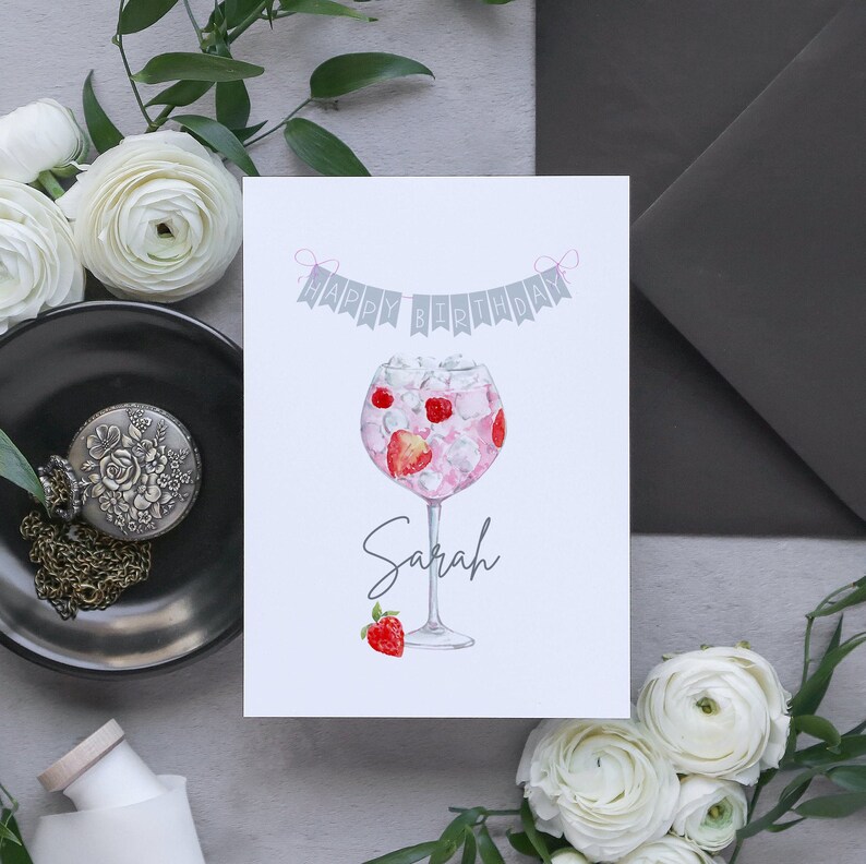 May include: A white card with a gray banner that says "Happy Birthday" with pink string. A watercolor illustration of a pink gin cocktail with strawberries and ice in a glass. The name "Sarah" is written below the glass.
