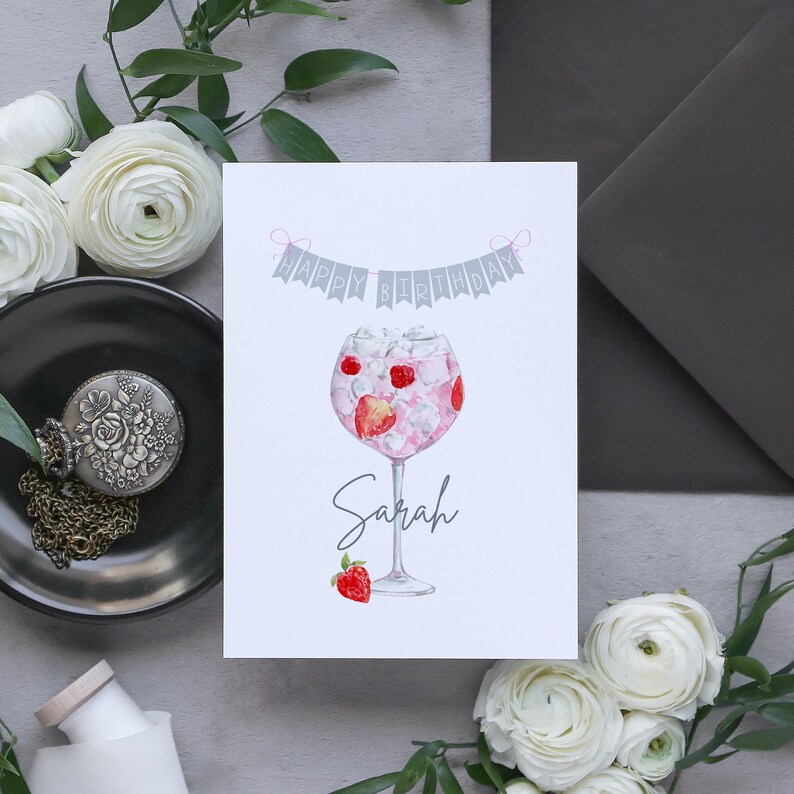 May include: A white card with a pink and gray happy birthday banner and a watercolor illustration of a pink gin cocktail with strawberries. The name Sarah is written below the cocktail.