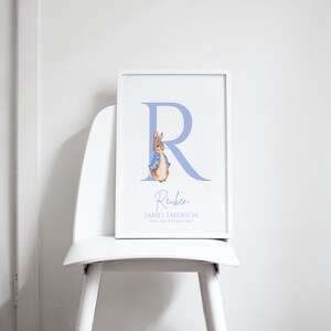 Pode incluir: Uma impressão de berçário personalizada azul e branca com um personagem de coelho com a letra "R" e o nome "Reuben James Emerson" com a data de nascimento "Nascido em 23 de dezembro de 2020".