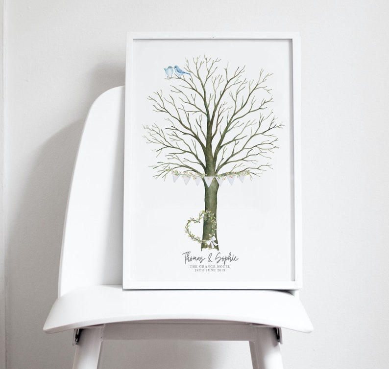 May include: A framed watercolor print featuring a tree with a heart-shaped wreath and a banner of small flags. Two blue birds perch atop the tree. The print includes names and a date, likely for a wedding or special event.