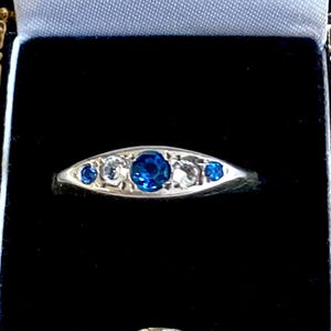 May include: An open black jewelry box with a gold trim, revealing a silver ring with a central blue gemstone and smaller clear stones. The ring is set against a black velvet interior, with a white satin lining. The box is on a gray surface.
