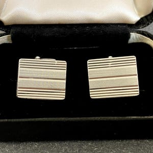 May include: A pair of rectangular silver-toned cufflinks featuring a horizontal striped pattern with alternating brushed and polished finishes. The cufflinks are presented inside a black hinged display box with white satin lining and the text JERMYN STREET S.W.1.