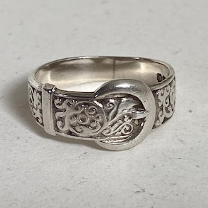 May include: A silver-toned metal ring designed to resemble a belt, featuring a buckle-shaped focal point and an embossed, intricate scrollwork pattern that wraps around the entire band, creating a continuous decorative motif across the surface.