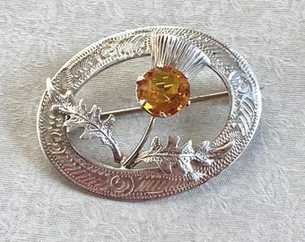 May include: Silver-toned brooch with an oval frame featuring a thistle design. The brooch has an amber-colored gemstone at the center and is secured with a pin. The brooch is likely a piece of jewelry.