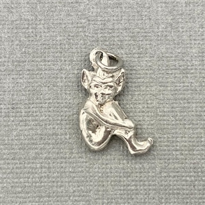 Puede incluir: Un dije de plata con forma de duende o elfo sentado. El dije tiene rasgos faciales detallados, orejas puntiagudas y está sosteniendo sus piernas. Tiene un lazo en la parte superior para sujetarlo a un collar o pulsera.