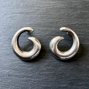 May include: A pair of silver-toned, curved earrings. Each earring has a smooth, polished finish and a spiral design. The earrings are displayed on a dark gray surface.