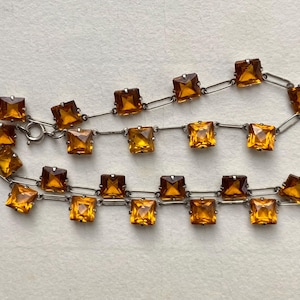 Könnte beinhalten: Eine Vintage-Halskette mit quadratischen, bernsteinfarbenen Glassteinen, die in einer silberfarbenen Metallkette gefasst sind. Die Halskette hat ein klassisches Design mit einem Verschluss. Die Steine wechseln sich in helleren und dunkleren Bernsteintönen ab.