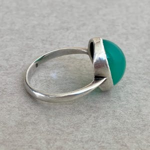 May include: A silver ring featuring a smooth, rounded, vibrant green cabochon stone set in a polished metal bezel. The band is thin and circular, showcasing a simple design that highlights the contrast between the bright green stone and the silver.