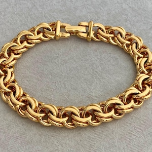 Puede incluir: Una pulsera de cadena en tono dorado que presenta una serie de eslabones redondeados entrelazados que crean un patrón denso y texturizado. La pulsera se asegura con un mecanismo de cierre de caja rectangular alineado con los eslabones.