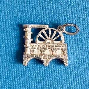May include: Silver charm shaped like a small building with a large wheel on the front. The charm has a loop at the top for attaching to a bracelet or necklace.