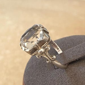 May include: A silver ring featuring a large, clear, rectangular-cut gemstone. The ring has an ornate setting with scroll detailing, and the word "SILVER" is stamped on the band. The gemstone is faceted, reflecting light.
