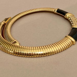 以下が含まれることがあります： リブ編みのデザインが施されたゴールドトーンのネックレス。黒いエナメルのアクセントと、小さな輝く石で装飾されたセクションが特徴です。留め金はゴールドトーンです。スタイリッシュなジュエリーです。