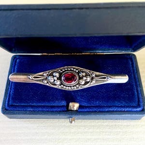 May include: A silver bar pin with an oval, red gemstone center, set in an ornate design. The pin is displayed in an open, blue jewelry box with a velvet lining. A classic piece of jewelry.
