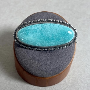 May include: Oval-shaped turquoise brooch with a silver-tone metal frame. The brooch features a textured, light blue stone set within a decorative border. The pin is displayed on a gray, rounded surface.