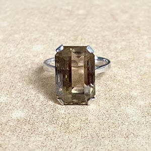 Puede incluir: Un anillo de plata con una gran gema rectangular de cuarzo ahumado. La gema es de color marrón translúcido y está engastada con cuatro pequeñas garras. La banda del anillo es de plata pulida.