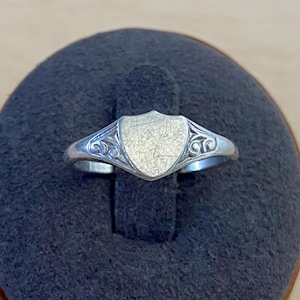 May include: A silver-toned ring featuring a central shield-shaped plaque with a brushed finish. The shoulders of the ring are decorated with intricate, raised scrollwork patterns that extend from the sides of the shield toward the band.