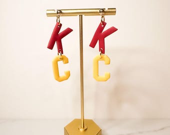 Puede incluir: Un par de soportes de pendientes de oro con abalorios de letras rojas y amarillas. Los abalorios deletrean "KC".