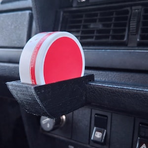 Può includere: Un accessorio per auto nero stampato in 3D contiene un contenitore bianco e rosso. Il supporto è montato sul cruscotto di un'auto, vicino alle bocchette dell'aria e al pannello di controllo. Il contenitore ha un coperchio circolare rosso.