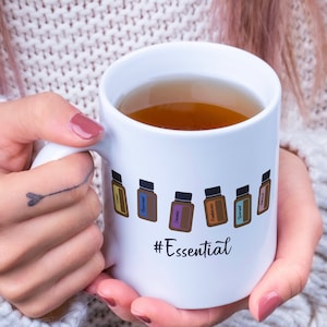 May include: White ceramic mug with a design of essential oil bottles and the text "#Essential". The mug is filled with a brown liquid and is being held by a person.