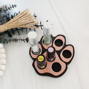 May include: A black and pink paw-shaped essential oil holder with six slots. The holder has a pink rim and is filled with five essential oil bottles. The bottles are labeled "Madagascar Vanilla" and "Rose".