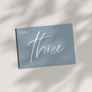 May include: A rectangular, light blue table number card with the word "TABLE" at the top left and the number "Three" in a white, cursive font. The card is angled, with shadows cast across it.