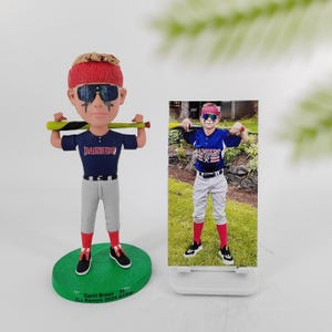 May include: A custom bobblehead figurine of a young baseball player wearing a blue and red jersey with the word "Rainiers" on it. The figurine is holding a baseball bat and is wearing sunglasses and a red headband. The figurine is standing on a green base with the text "Gavin Brown, 33, CLL Rainiers 2024 All-Star" printed on it. A photo of the same player in a baseball uniform is displayed on a white stand behind the figurine.