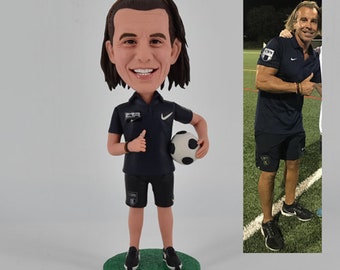 May include: A custom bobblehead figurine of a person wearing a black soccer jersey with a white logo and black shorts. The figurine is holding a soccer ball and giving a thumbs up. The figurine is standing on a green base.