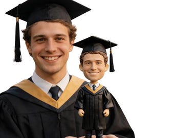 May include: A smiling graduate in a black cap and gown holds a bobblehead figurine of himself, also in graduation attire. The bobblehead has a black cap, a black gown, and a gold trim.