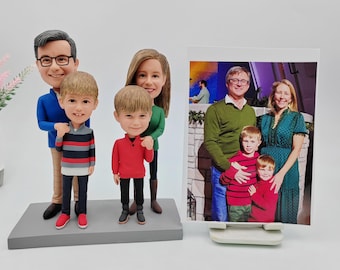May include: A family of five custom-made figurines with a photo of the family in a frame. The figurines are made of painted ceramic and are dressed in casual clothing. The photo shows the family standing together in front of a stage.