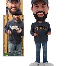 May include: A custom bobblehead figurine of a person wearing a blue hoodie with the word "Michigan" on it, a blue baseball cap with a yellow "M" on it, and blue jeans. The person is holding a bottle of beer in their right hand.
