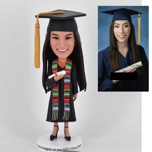 Könnte beinhalten: Eine Bobblehead-Figur mit Abschlusscap und -robe mit einem bunten Schal. Die Figur hält ein Diplom. Der Bobblehead steht auf einem Sockel mit dem Text "Herzlichen Glückwunsch zu deinem Abschluss 2022".