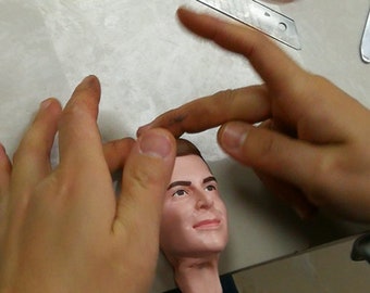 May include: A person's hands are carefully working on a sculpted human head, which is made of a light brown material. The head has realistic features, including brown hair, brown eyes, and a painted face.