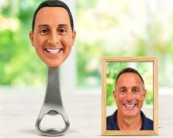 May include: A custom bottle opener featuring a three-dimensional sculpted head of a smiling person with short dark hair, attached to a silver metal handle. Beside it stands a rectangular wooden picture frame containing a photograph of the same person.
