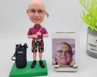 May include: A custom caricature figurine of a person wearing a pink golf shirt, colorful shorts, and holding a golf club. The figurine is standing on a green base with a black golf bag. A photo of the person is displayed on a stand next to the figurine.
