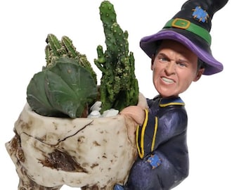 May include: A novelty planter featuring a skull-shaped pot filled with cacti and succulents. The planter is held by a figurine of a man in a wizard costume and a black witch hat with a green buckle. The figurine has an angry expression.