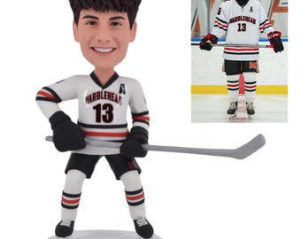 May include: A custom bobblehead of a hockey player wearing a white jersey with black and red trim. The jersey has the text "Marblehead" and the number "13" on the front. The bobblehead is holding a hockey stick.