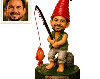 May include: A custom bobblehead figurine featuring a man with a beard wearing a red pointed hat, a green vest, and light-colored pants. The figure holds a black fishing rod with a red line and an orange fish, standing on a base labeled GONE FISHIN'.
