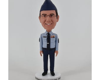 May include: A bobblehead figurine of a man in a light blue Air Force uniform, complete with a dark blue cap and tie. The figurine has a smiling face and glasses. The base of the figurine has text that reads "David Steven Geczy - USAF Retired - 1968-1994".