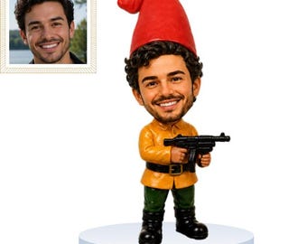 May include: A custom bobblehead figurine featuring a smiling man with dark curly hair and a beard, wearing a bright red gnome hat, a yellow collared shirt, a black belt, green pants, and black boots, while holding a black toy submachine gun.