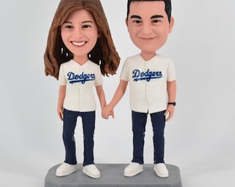 May include: A custom bobblehead couple wearing white baseball jerseys with the word "Dodgers" in blue. The couple is holding hands and standing on a gray base with the text "John and Lorena".